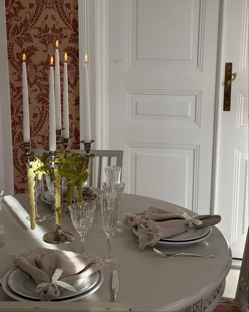 Juhlapöydän kattaus jossa näkyy pellavaiset vuokrattavat servetit. Vanhan valkoisen pöydän päällä hopeinen kynttelikkö johon on kiedottu revonhäntää, hopeoidut aterimet, kristalliviinilasit, valkoiset ruokalautaset ja kangasservetit.